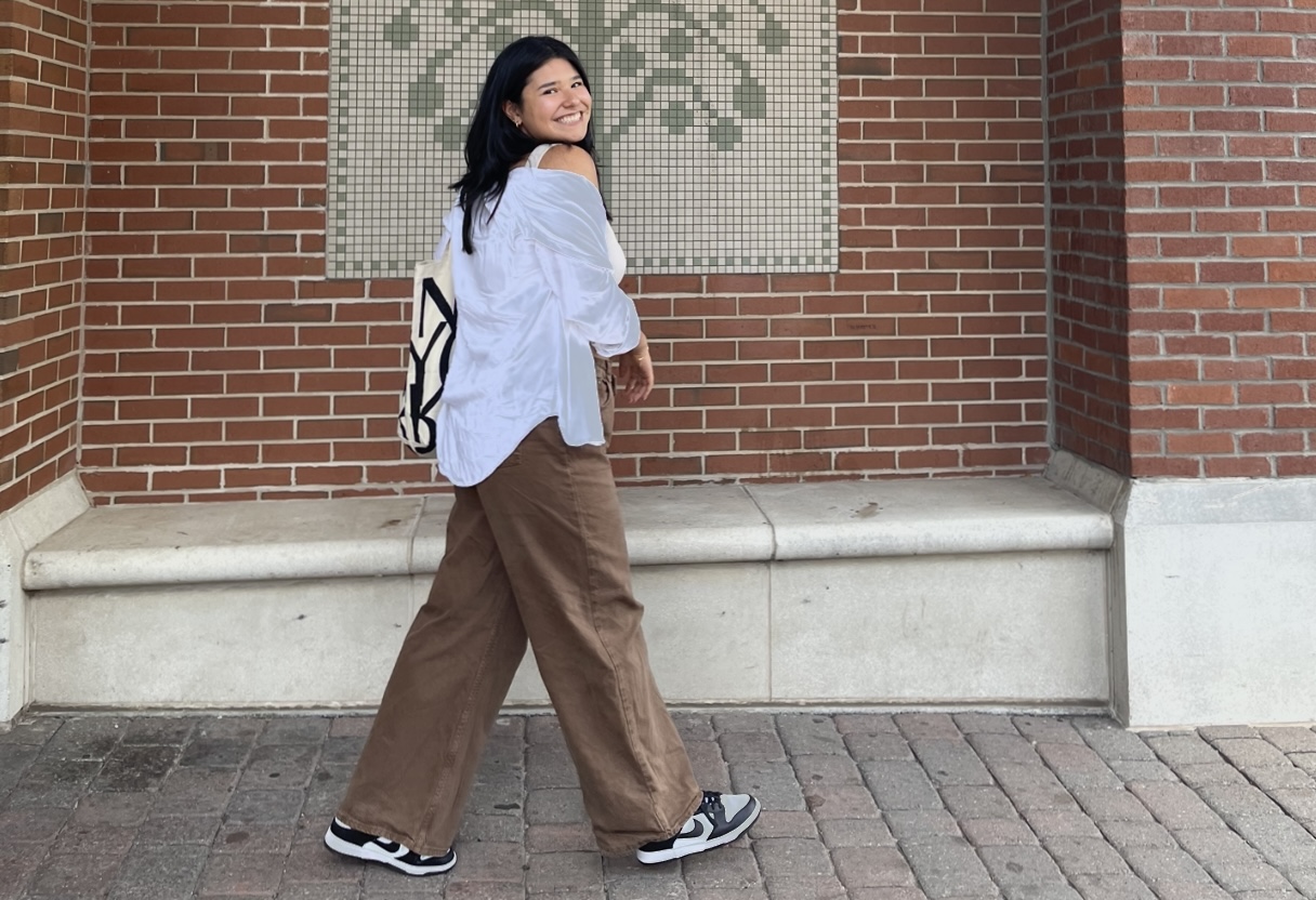 Picture of Megan wearing a white top, brown pants, and sneakers. She's smiling and walking past the camera, mid-stride.